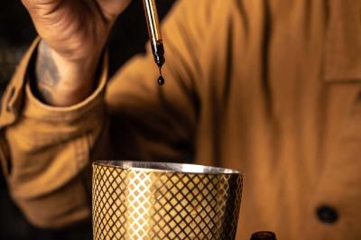 Un barman verse un liquide sombre d'une petite bouteille dans un verre à cocktail rempli d'une boisson mousseuse.