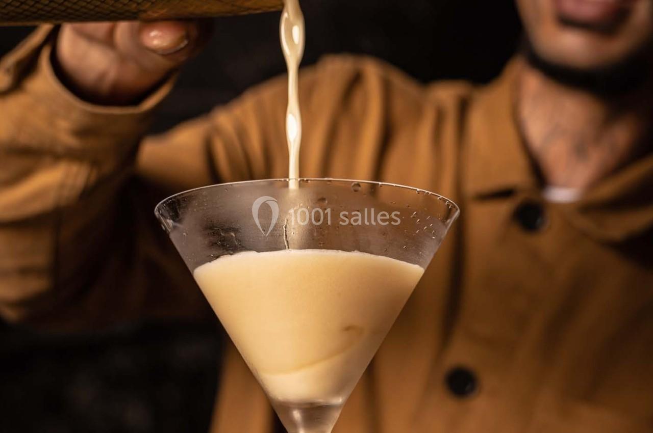 Un liquide beige est versé dans un verre à cocktail en forme de cône, tenu par une personne en veste marron.