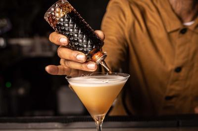 Un barman verse un liquide sombre d'une petite bouteille dans un verre à cocktail rempli d'une boisson mousseuse.
