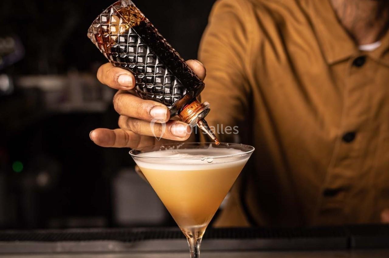 Un barman verse un liquide sombre d'une petite bouteille dans un verre à cocktail rempli d'une boisson mousseuse.