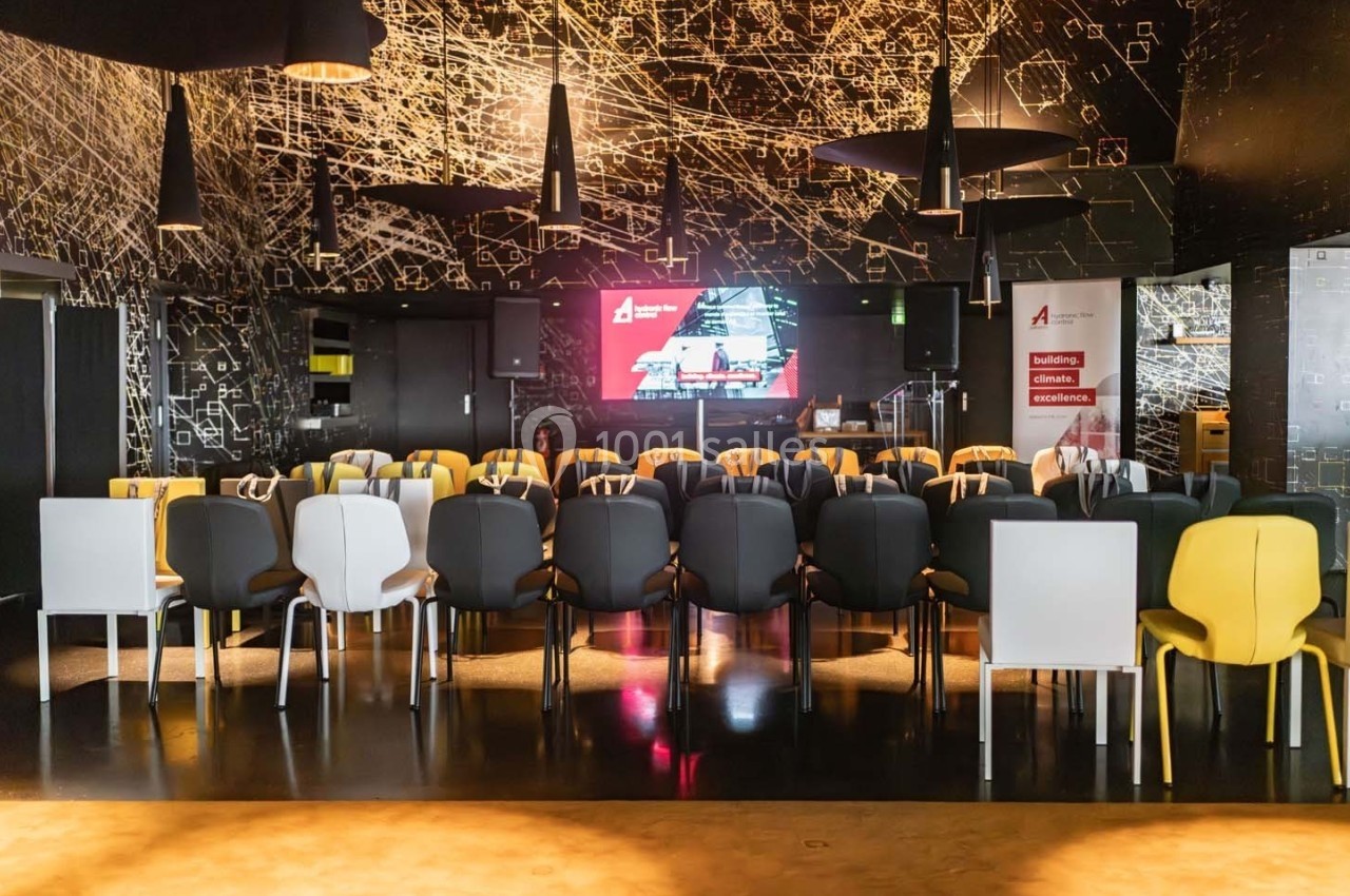 Salle de conférence moderne avec des rangées de chaises colorées face à un écran et un décor mural sombre.