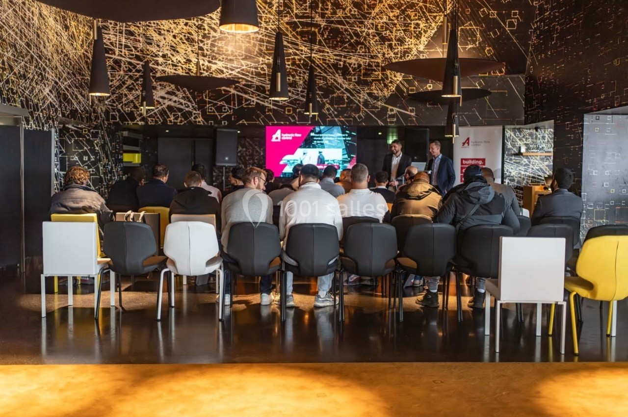 Groupe de personnes assises dans une salle de conférence moderne, écoutant une présentation sur un écran.