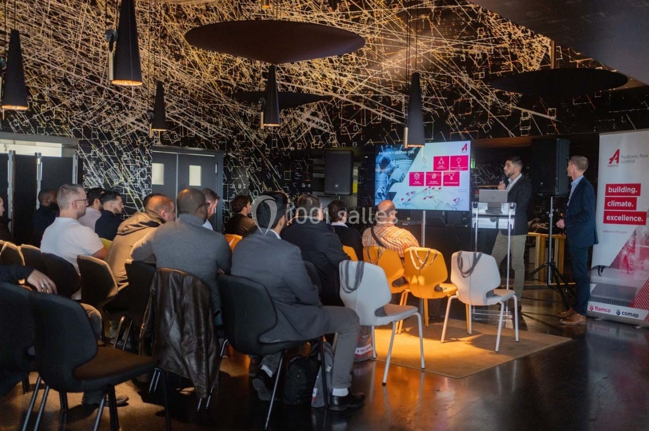 Un conférencier présente un diaporama à un public assis dans une salle moderne avec un plafond décoratif.