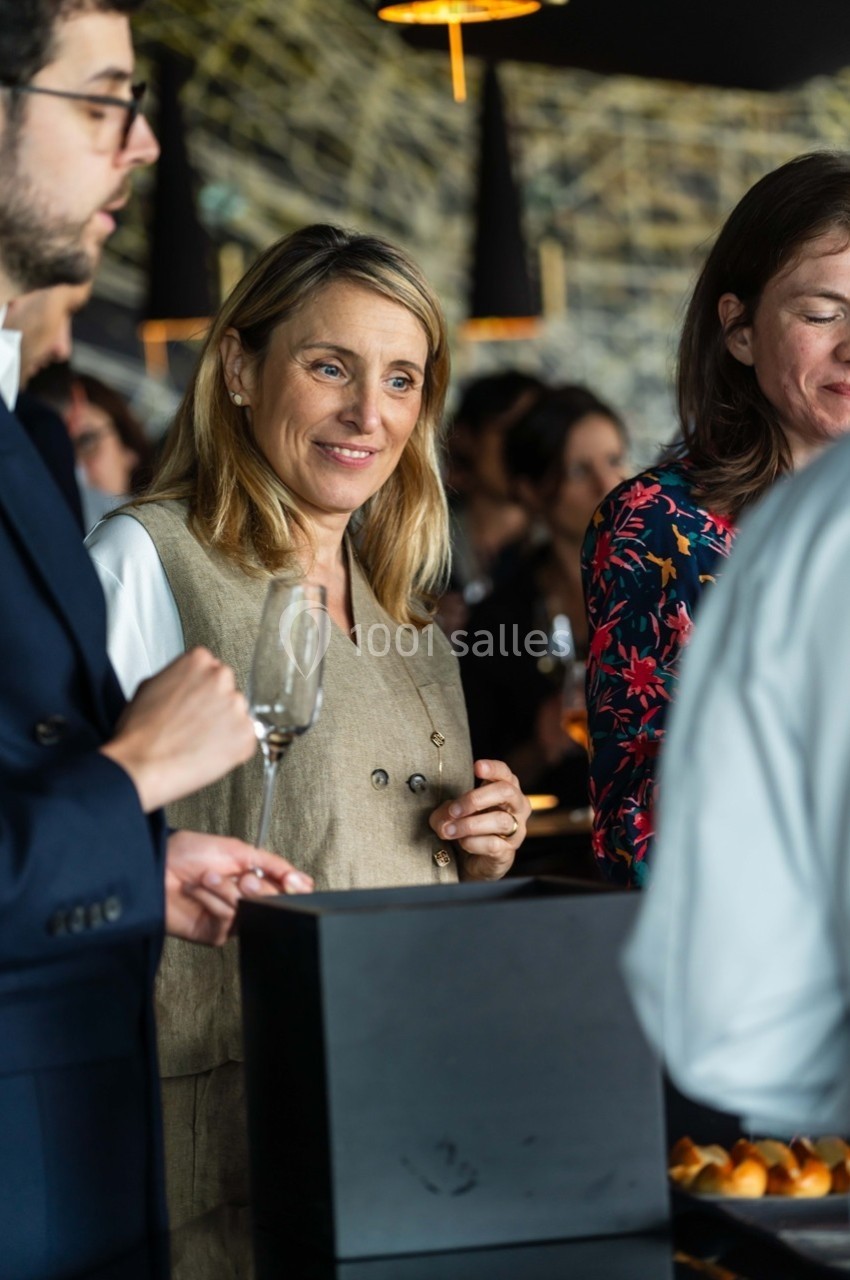 Une femme souriante tient un verre dans un cadre convivial avec d'autres personnes en arrière-plan.