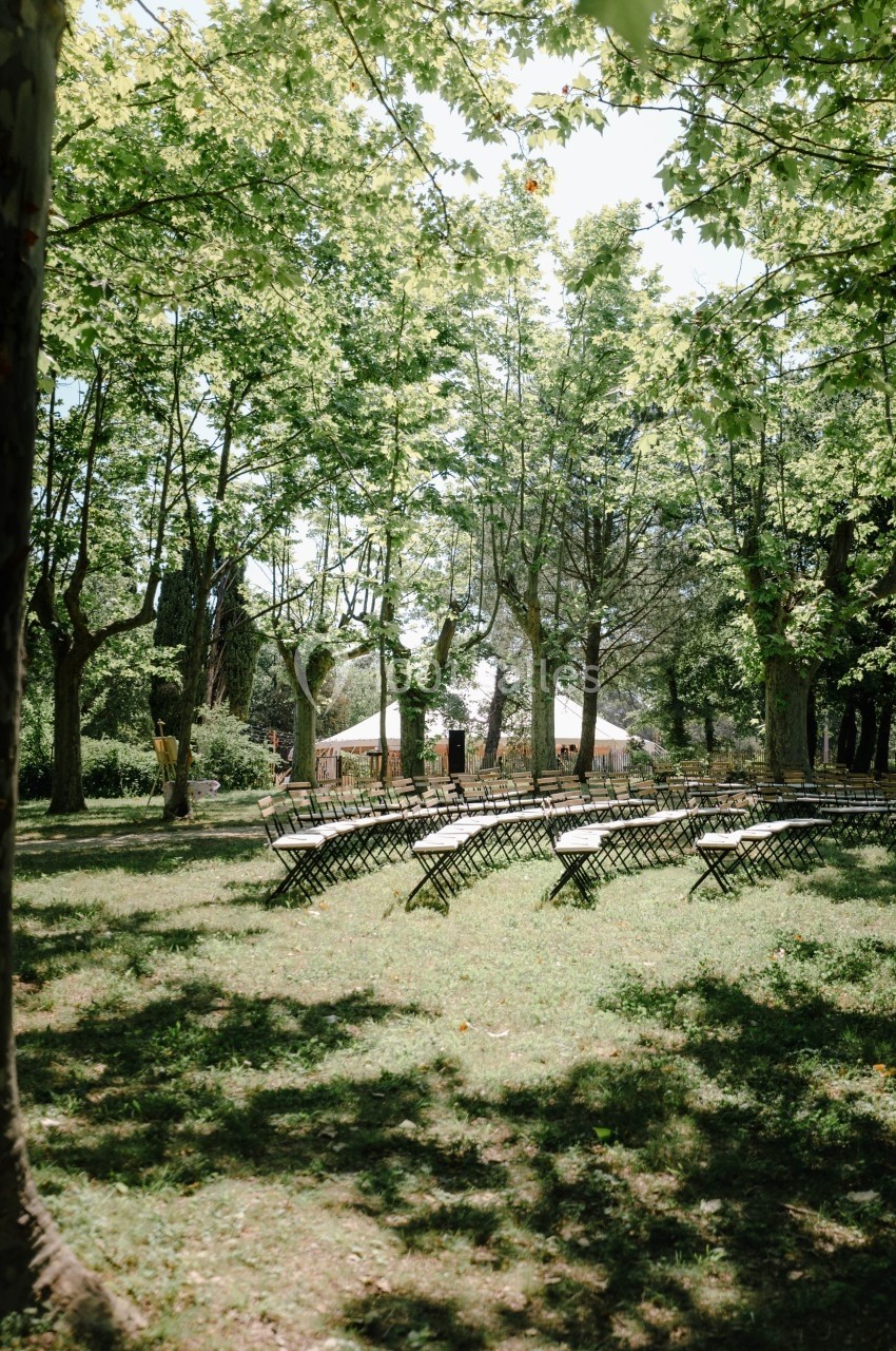Chaises disposées en arc de cercle sous des arbres dans un parc, avec une tente blanche en arrière-plan.