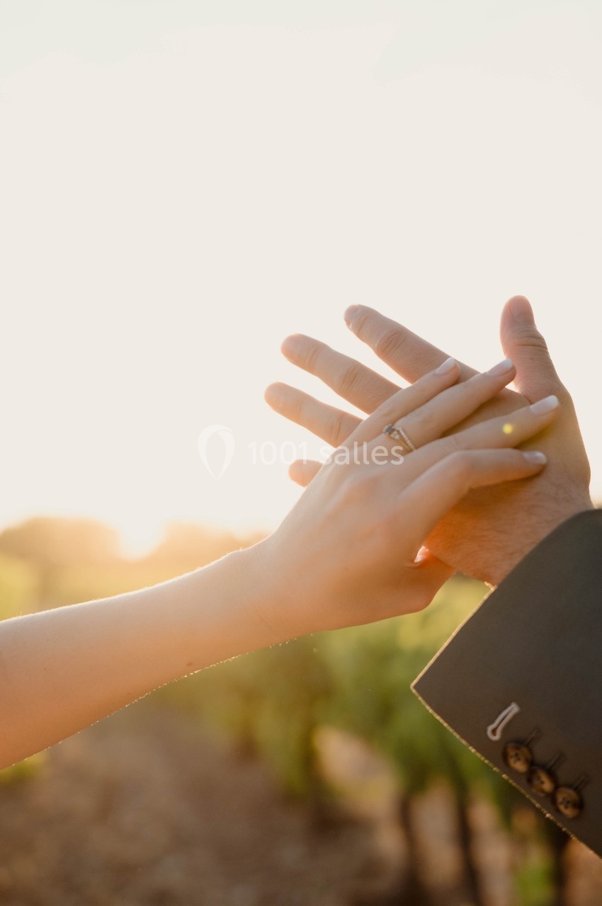 Deux mains entrelacées devant un coucher de soleil, l'une portant une alliance, dans un cadre naturel.