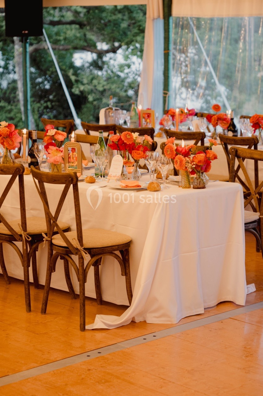 Table décorée avec nappes blanches, chaises en bois et bouquets de fleurs orange dans une salle lumineuse.