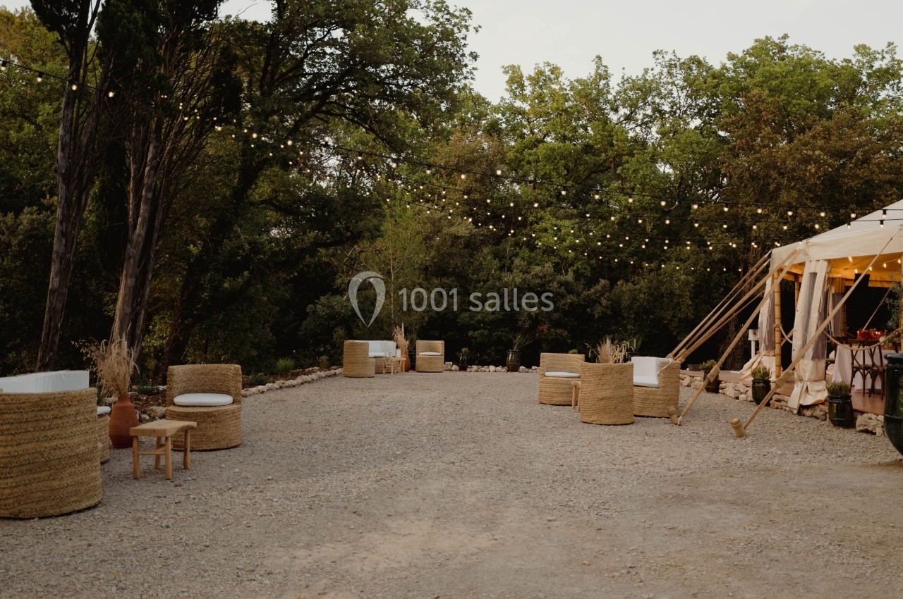 Espace extérieur avec fauteuils en rotin, guirlandes lumineuses et tente blanche dans un cadre arboré.