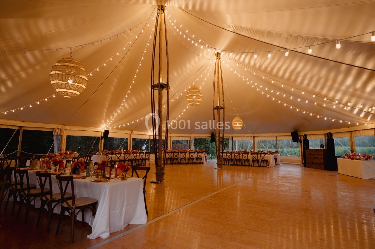 Intérieur d'une tente de réception éclairée par des guirlandes lumineuses, avec tables dressées pour un événement.