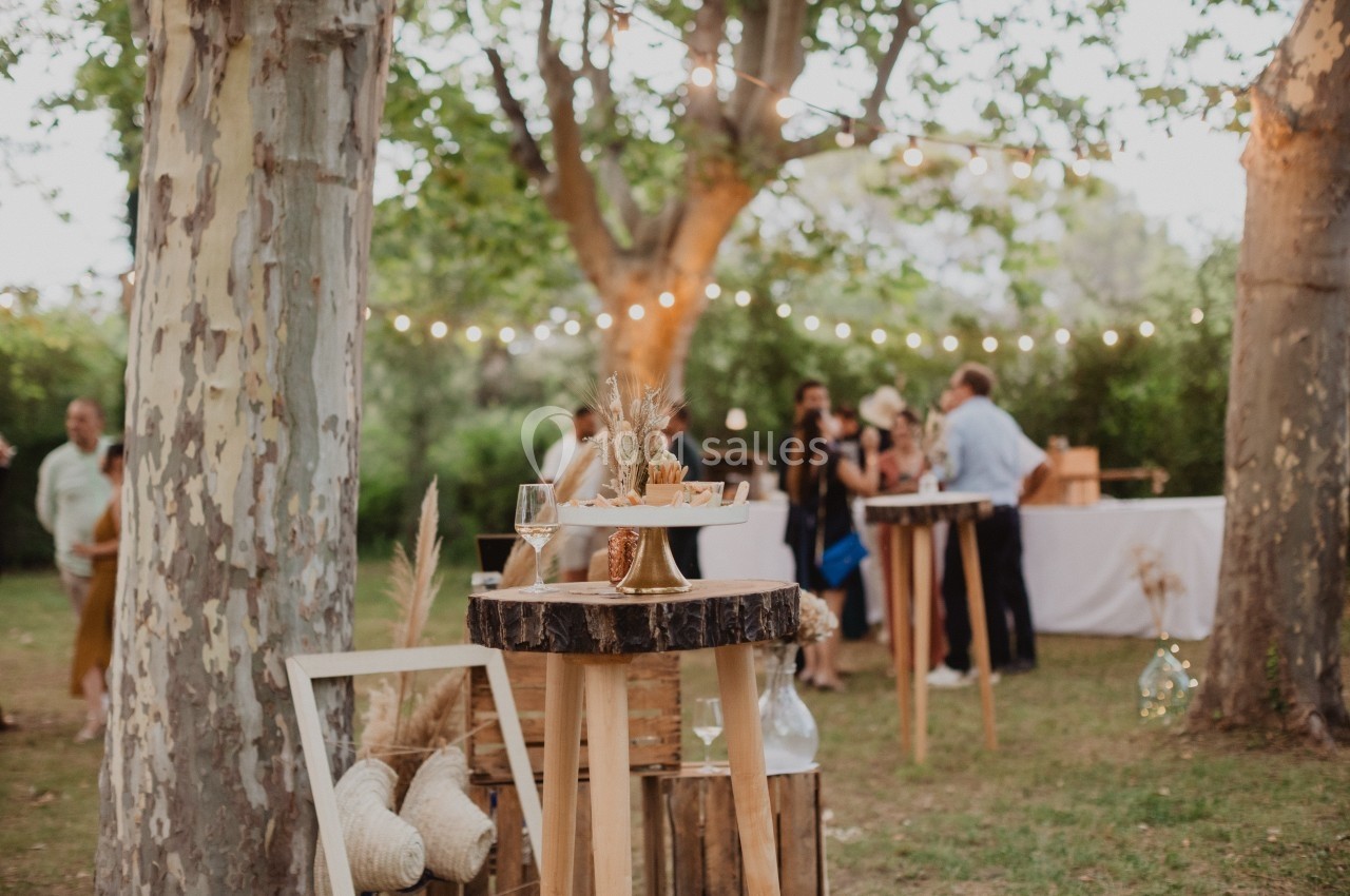 Tables en bois décorées avec des éléments naturels dans un jardin, avec des invités en arrière-plan sous des guirlandes…