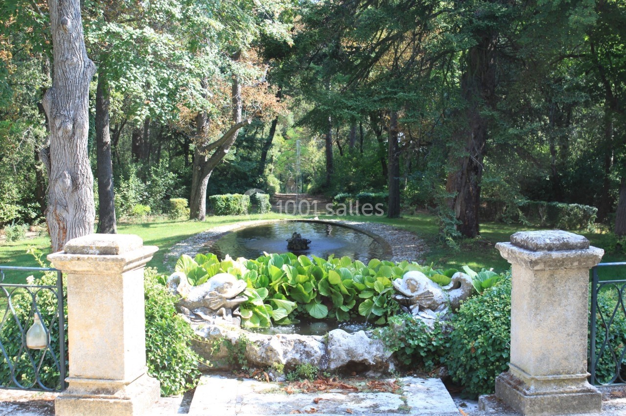 Vue d'un jardin avec un bassin entouré de végétation, des statues de cygnes et des arbres en arrière-plan.
