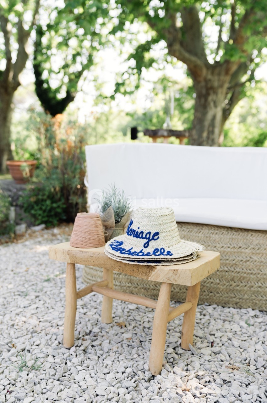 Chapeau en paille brodé posé sur un tabouret en bois, entouré de plantes et d'un canapé extérieur sous des arbres.