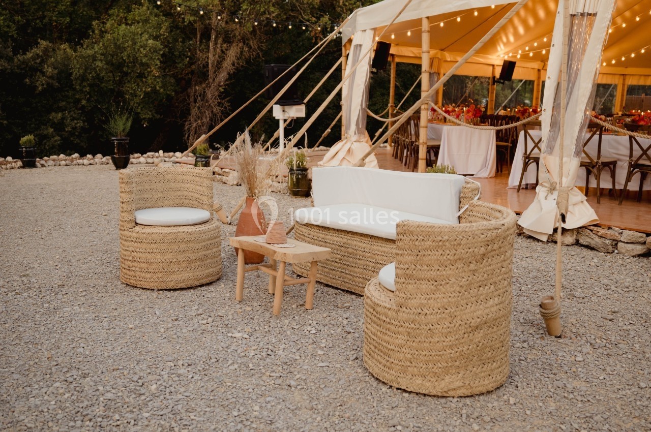 Mobilier en rotin avec coussins blancs disposé sur du gravier devant une tente éclairée pour un événement en extérieur.