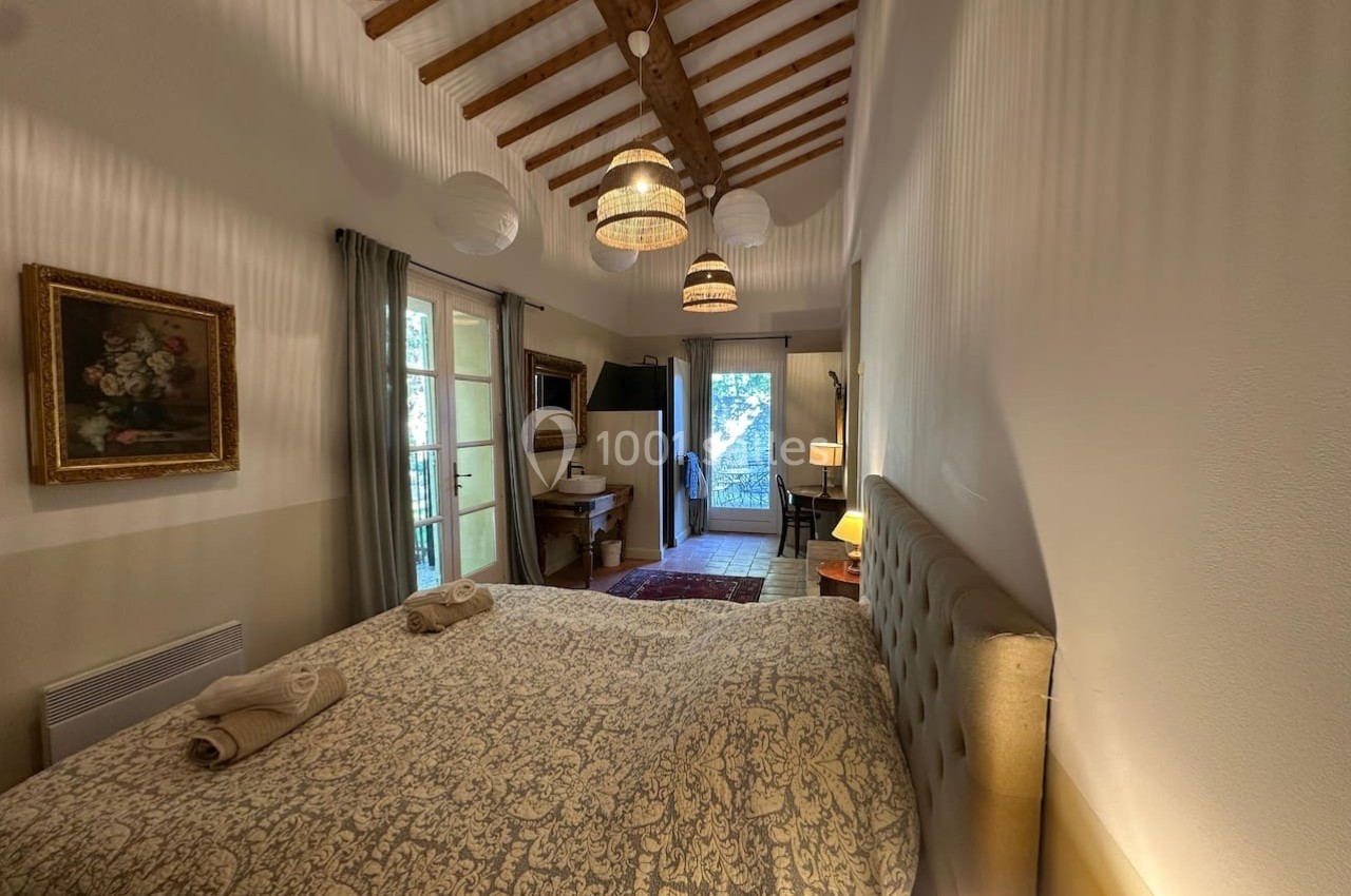 Chambre lumineuse avec lit double, poutres apparentes, décoration classique et vue sur un balcon arboré.