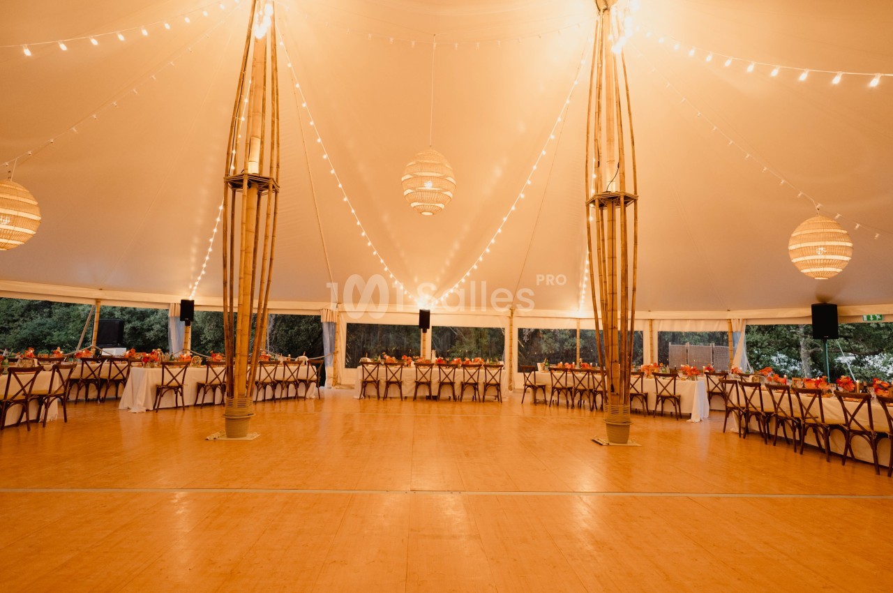 Salle de réception sous une tente éclairée avec des guirlandes lumineuses, tables décorées et espace central dégagé.