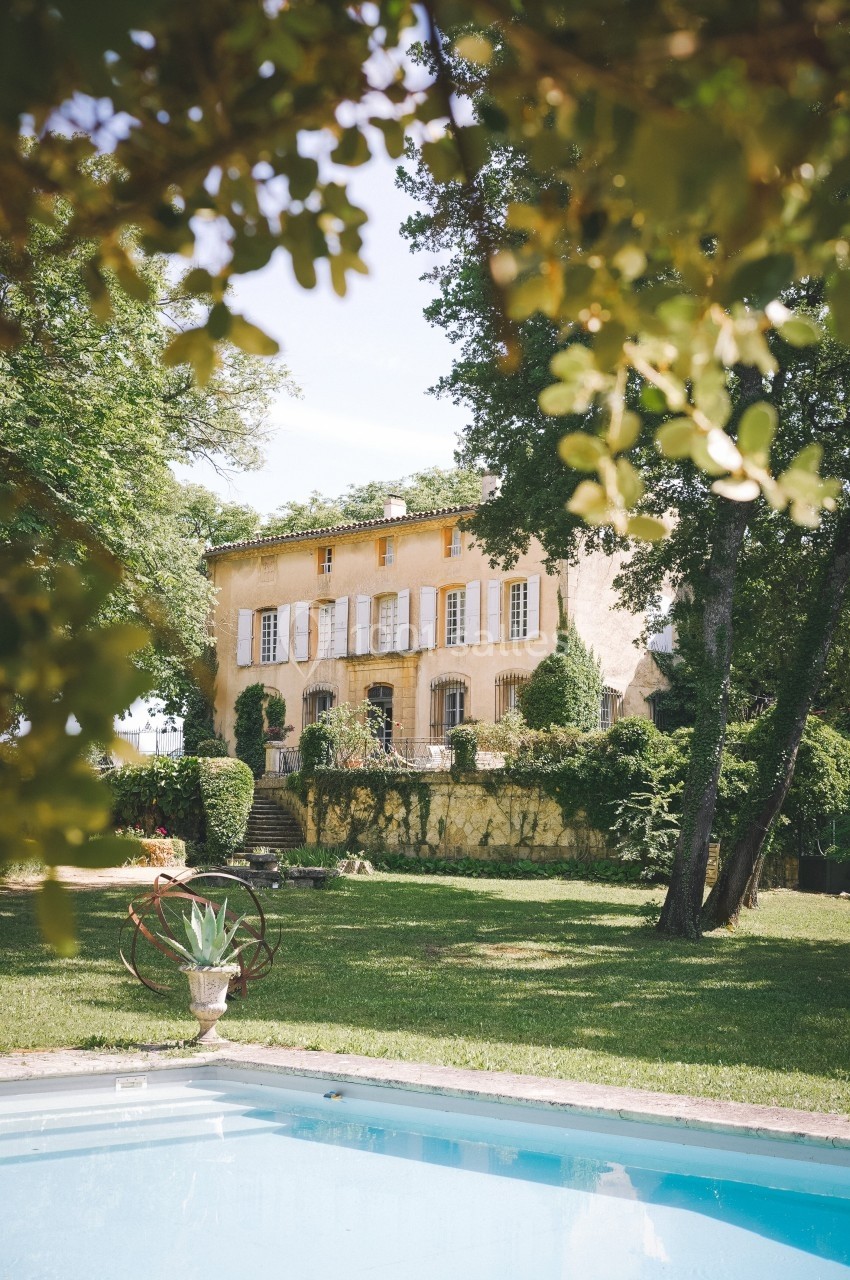 Grande maison en pierre entourée de verdure, avec une piscine au premier plan et des arbres en arrière-plan.