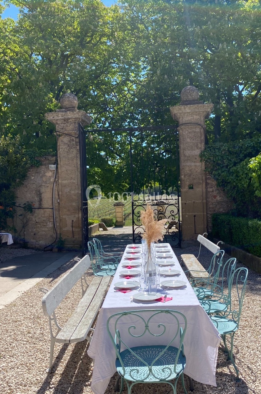 Table dressée en extérieur avec nappes blanches, chaises en métal, décor champêtre et portail ouvert sur un jardin.