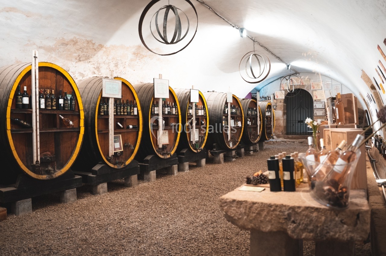 Cave voûtée avec des tonneaux en bois contenant des bouteilles de vin, et une table avec des produits locaux au premier plan.