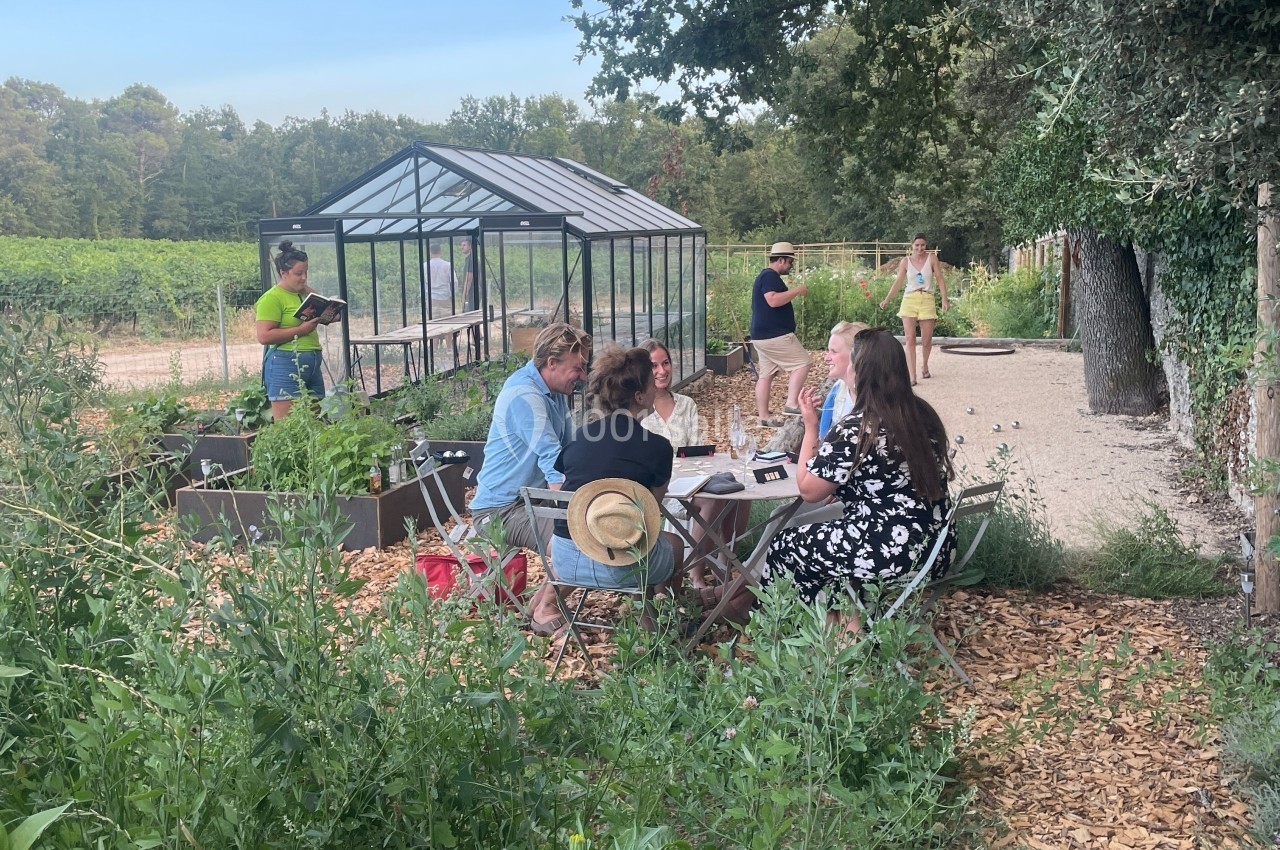 Un groupe de personnes discute autour d'une table en plein air près d'une serre, entouré de verdure.