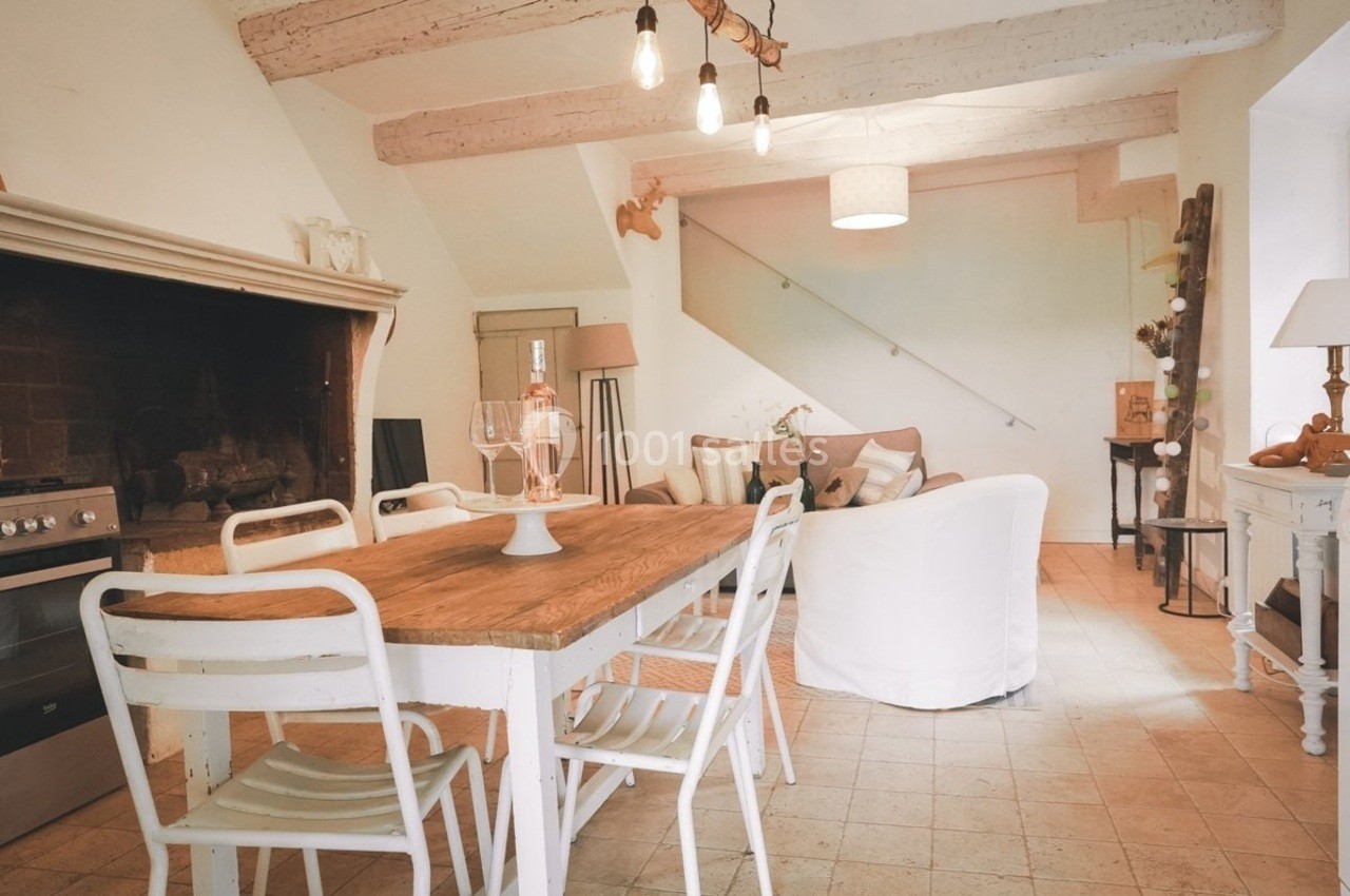 Salle à manger rustique avec table en bois, chaises blanches, cheminée ancienne et salon en arrière-plan.