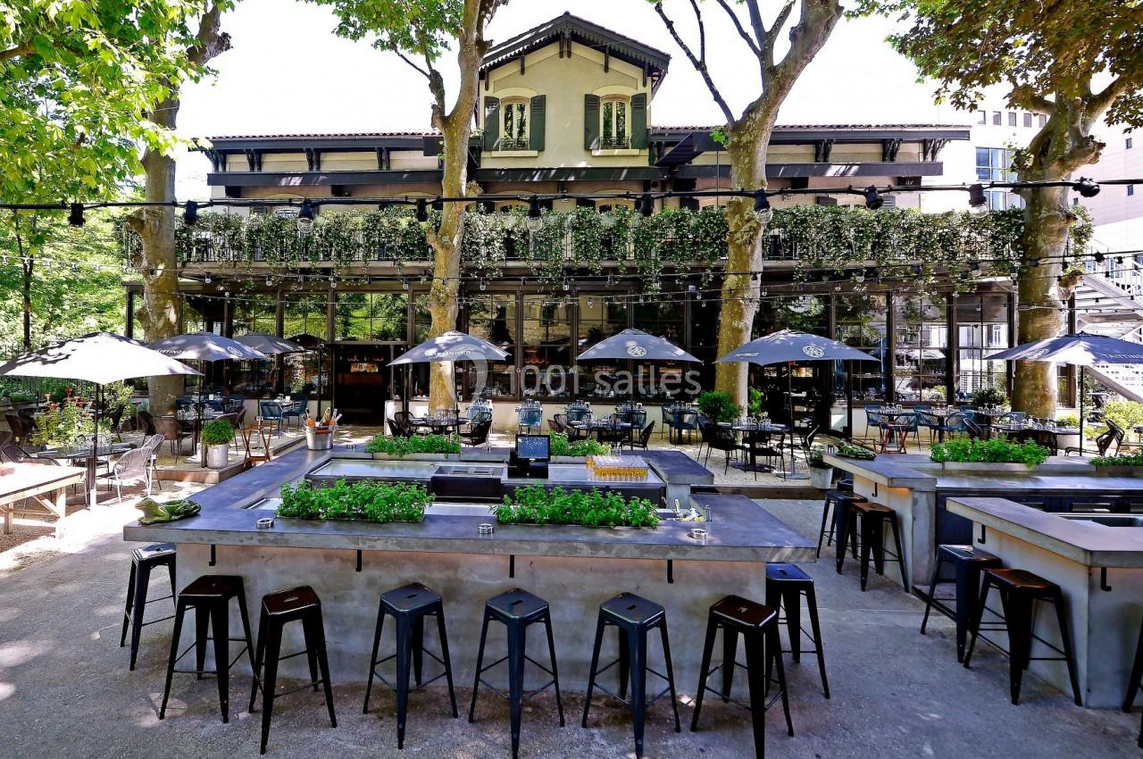 Terrasse extérieure d'un restaurant avec tables, chaises et bar, entourée d'arbres et d'une façade végétalisée.