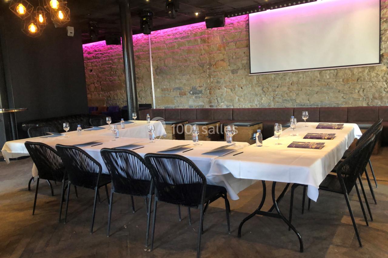 Salle de réunion avec tables dressées, chaises noires, murs en pierre et éclairage tamisé.