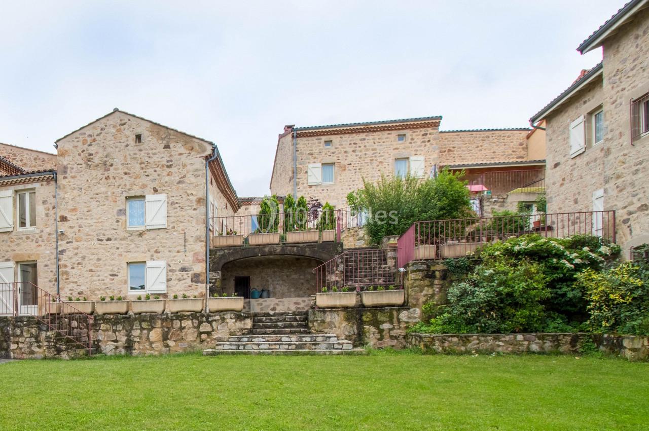 Bâtiments en pierre avec volets blancs entourant une cour verdoyante et des escaliers en pierre.