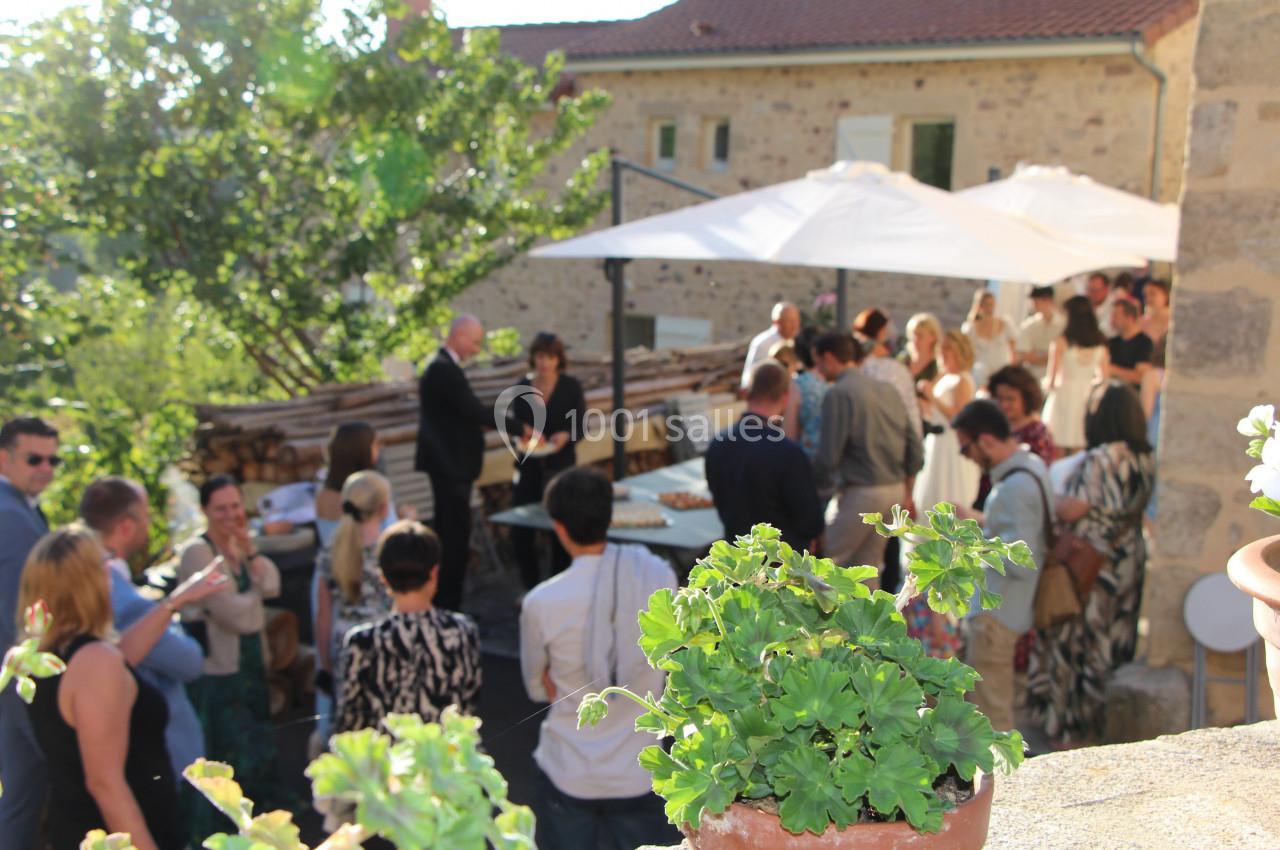 Groupe de personnes réunies en extérieur autour de tables, avec des plantes et un bâtiment en pierre en arrière-plan.