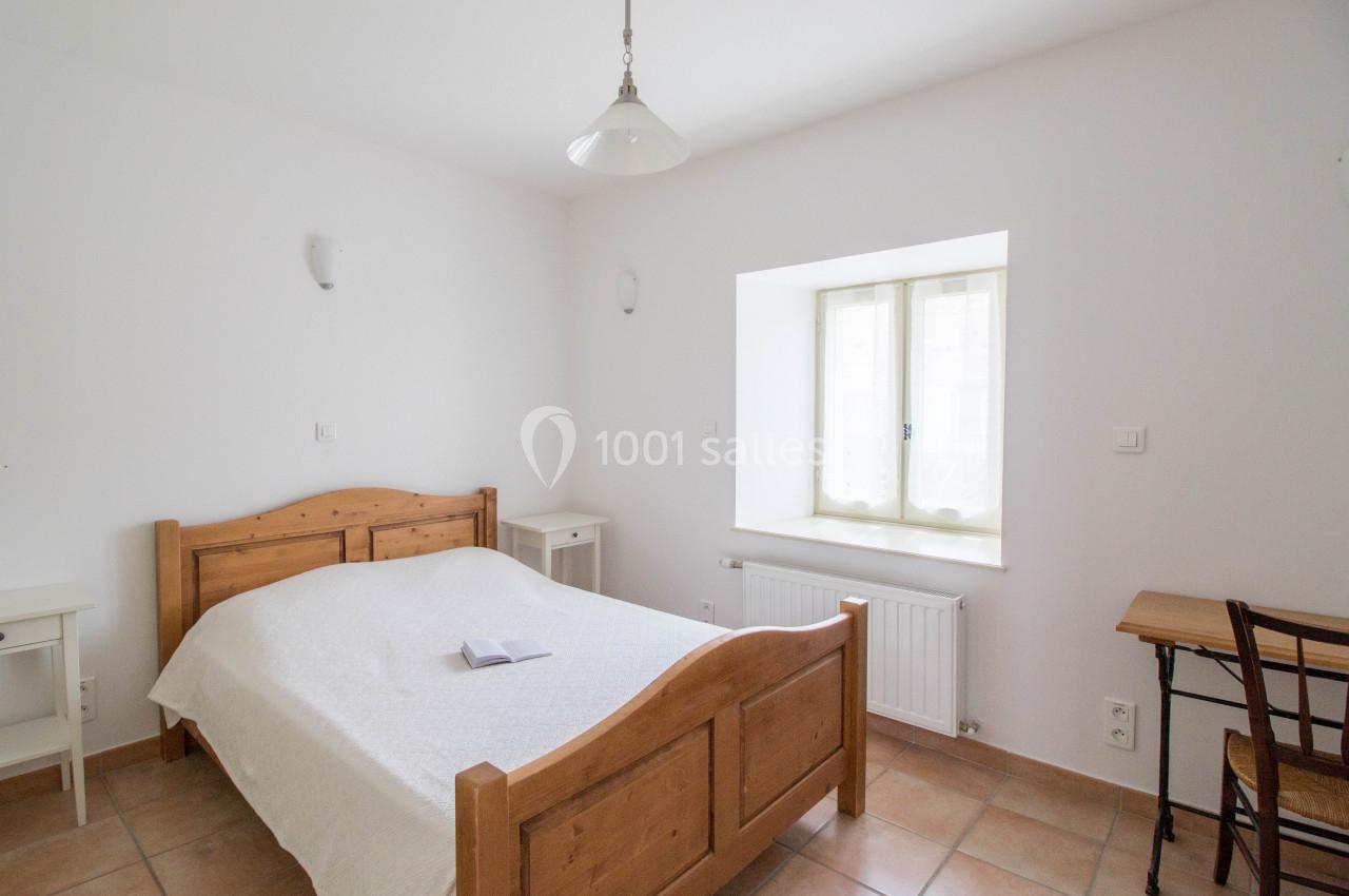 Chambre lumineuse avec un lit en bois, une table, une chaise, et une fenêtre laissant entrer la lumière naturelle.