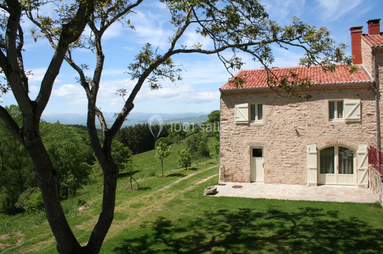 Maison en pierre avec toit en tuiles rouges, entourée de verdure et d'arbres, sous un ciel bleu dégagé.