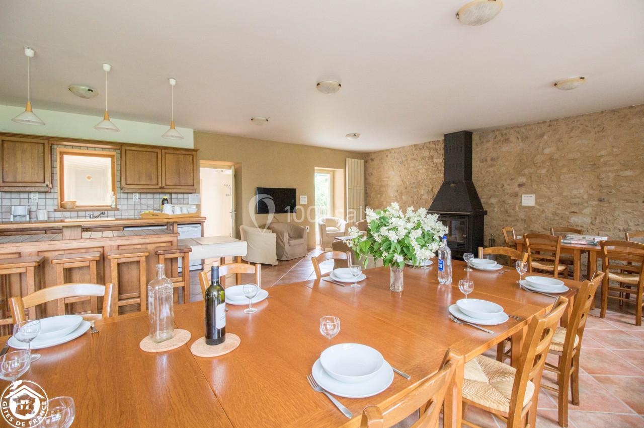 Salle à manger lumineuse avec une grande table en bois, cheminée, cuisine ouverte et murs en pierre apparente.