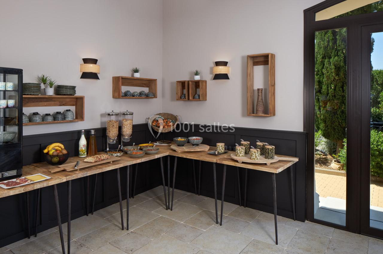 Buffet de petit-déjeuner avec céréales, fruits, pain et boissons, disposé sur des tables en bois dans une salle lumineuse.