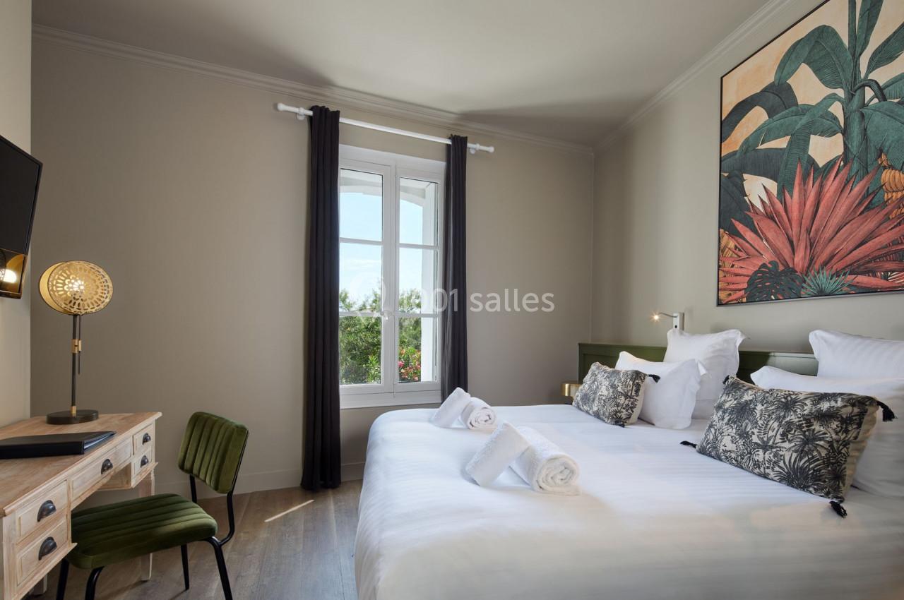 Chambre lumineuse avec lit double, bureau en bois, chaise verte, grande fenêtre et décoration murale colorée.