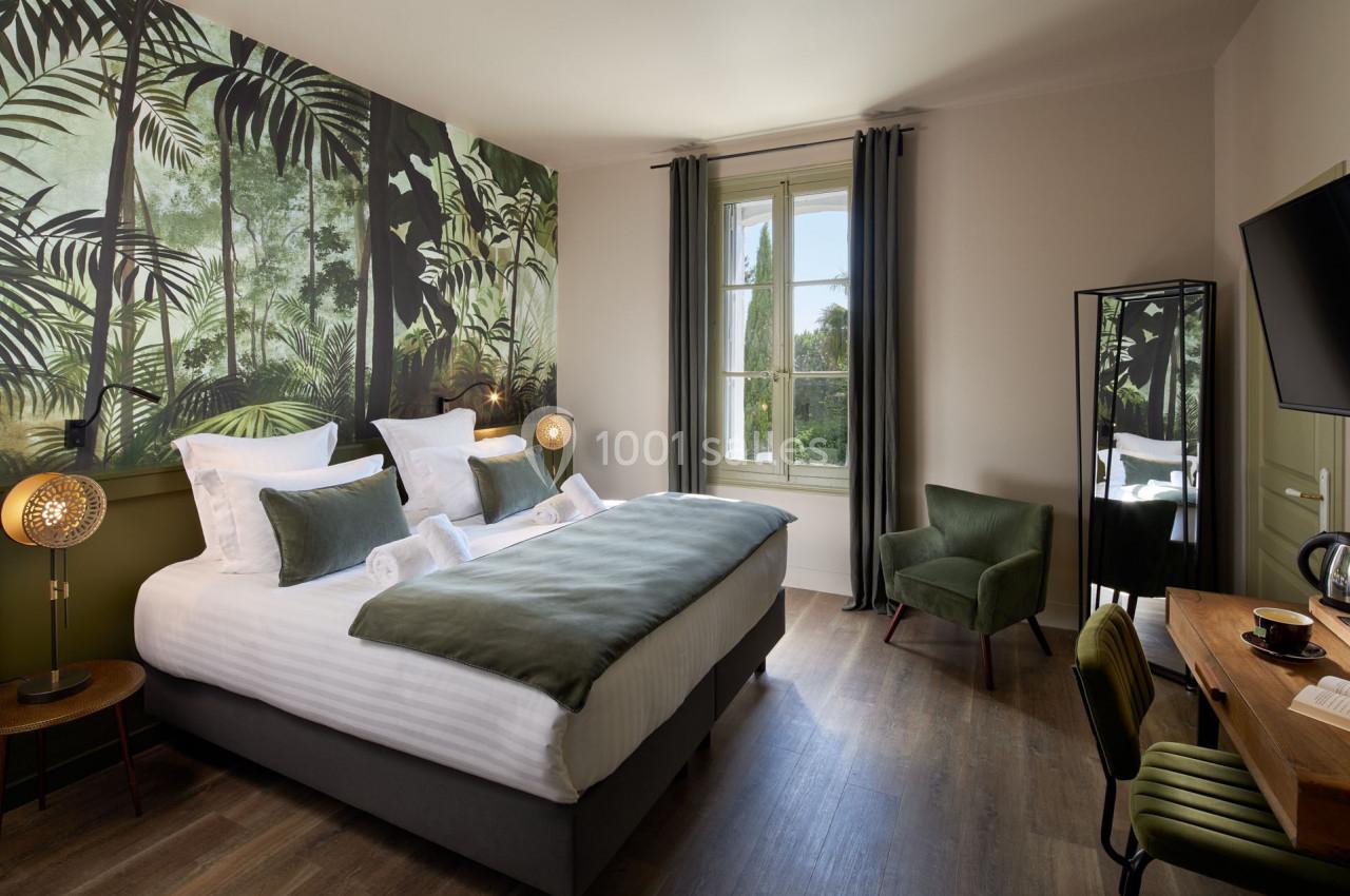 Chambre lumineuse avec un lit double, décoration murale tropicale, fauteuil vert, bureau et grande fenêtre.