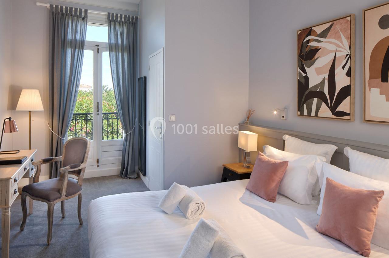 Chambre lumineuse avec lit double, coussins roses, bureau, chaise, lampes et fenêtre donnant sur un balcon.