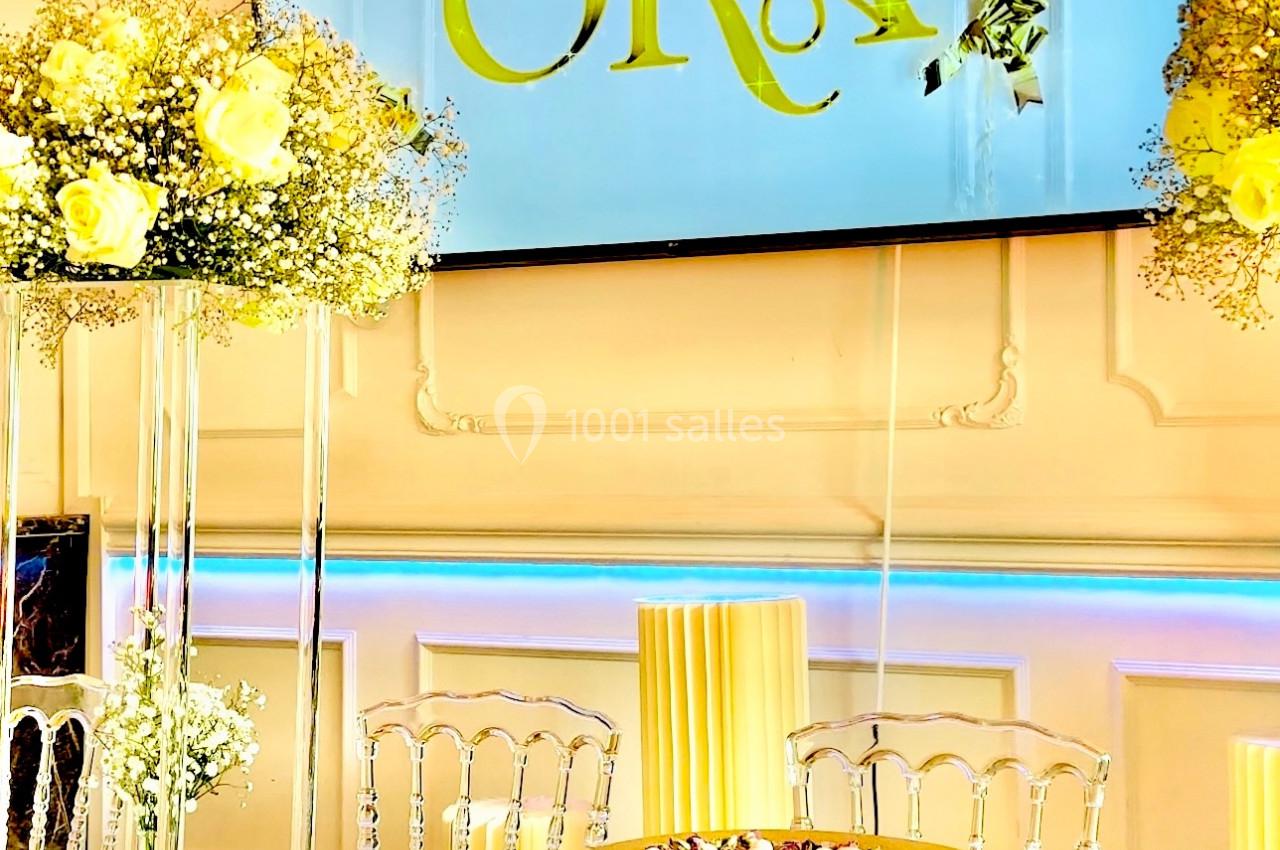 Salle décorée avec des chaises transparentes, une table ornée de fleurs et un écran affichant ’La Villa Ora’.