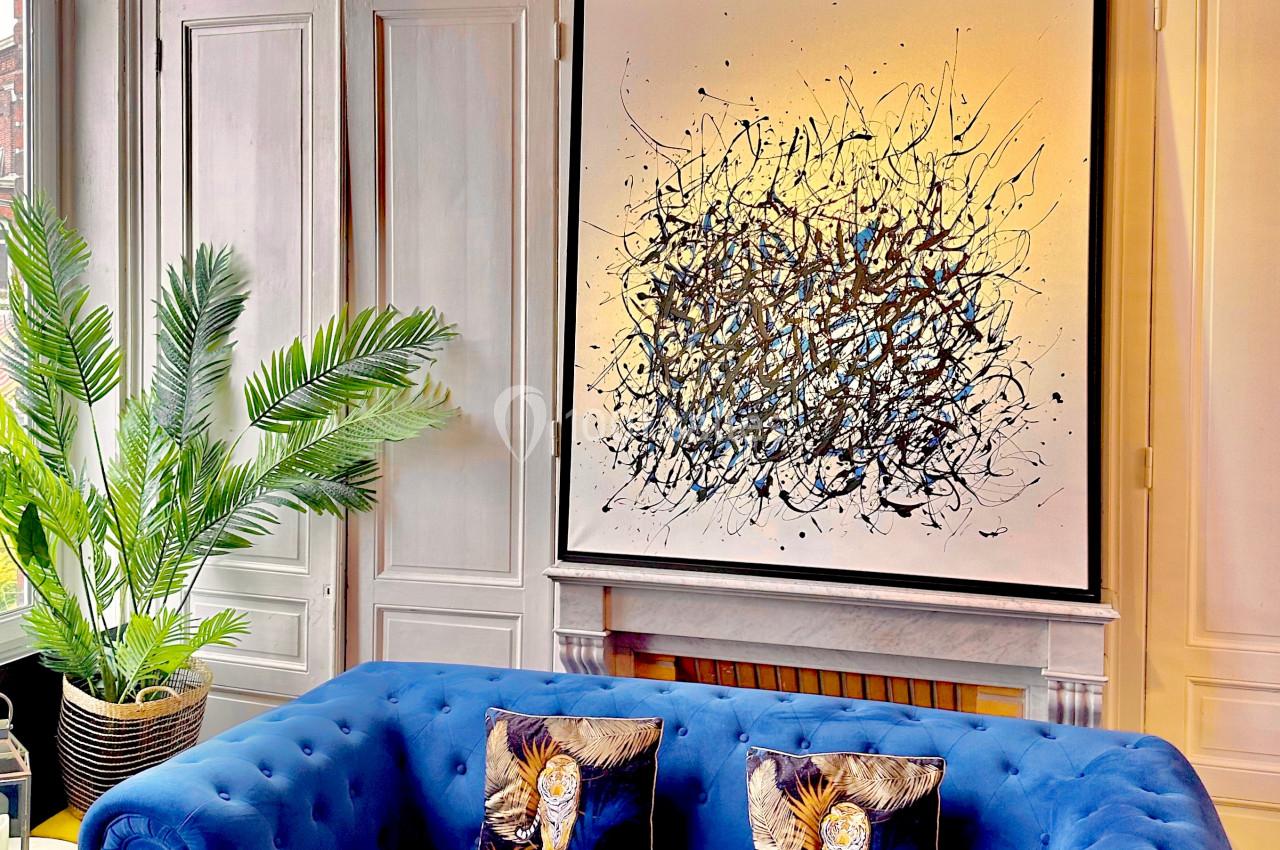 Canapé bleu avec coussins décoratifs, tableau abstrait au mur, plante verte et lampe suspendue dans un salon lumineux.