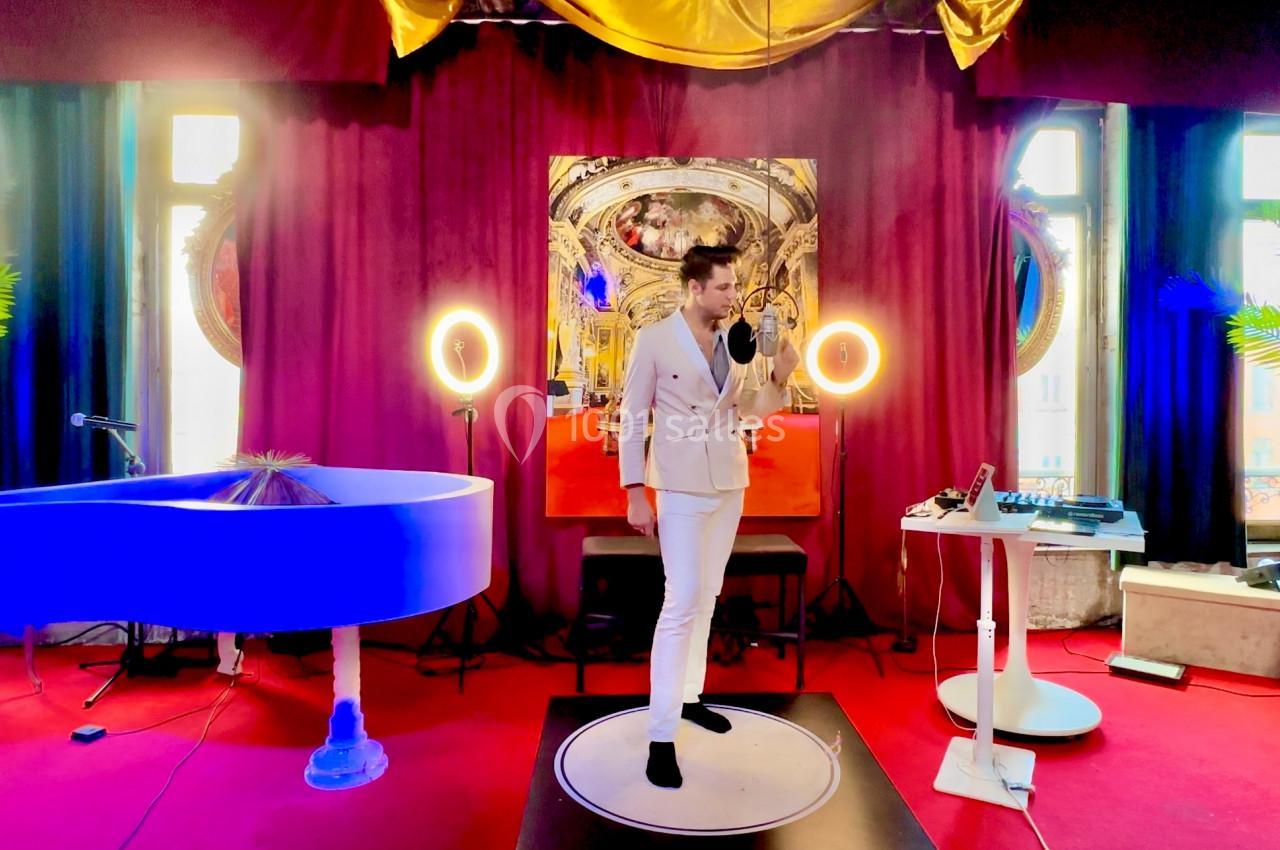 Une personne en costume blanc chante sur une scène avec rideaux rouges, piano bleu et éclairage circulaire.