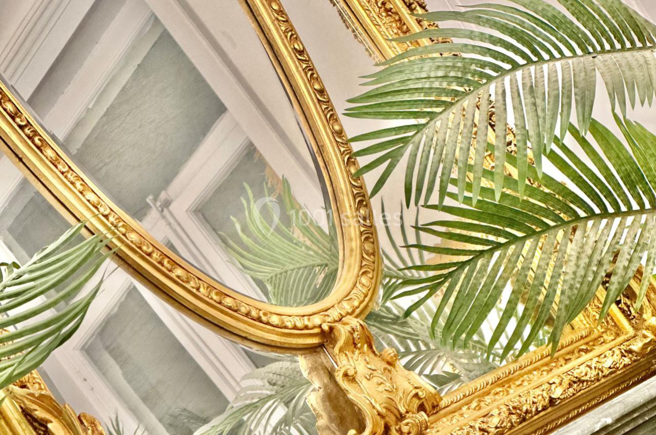 Miroir doré orné de détails sculptés, reflétant une fenêtre, entouré de feuilles de plantes vertes.