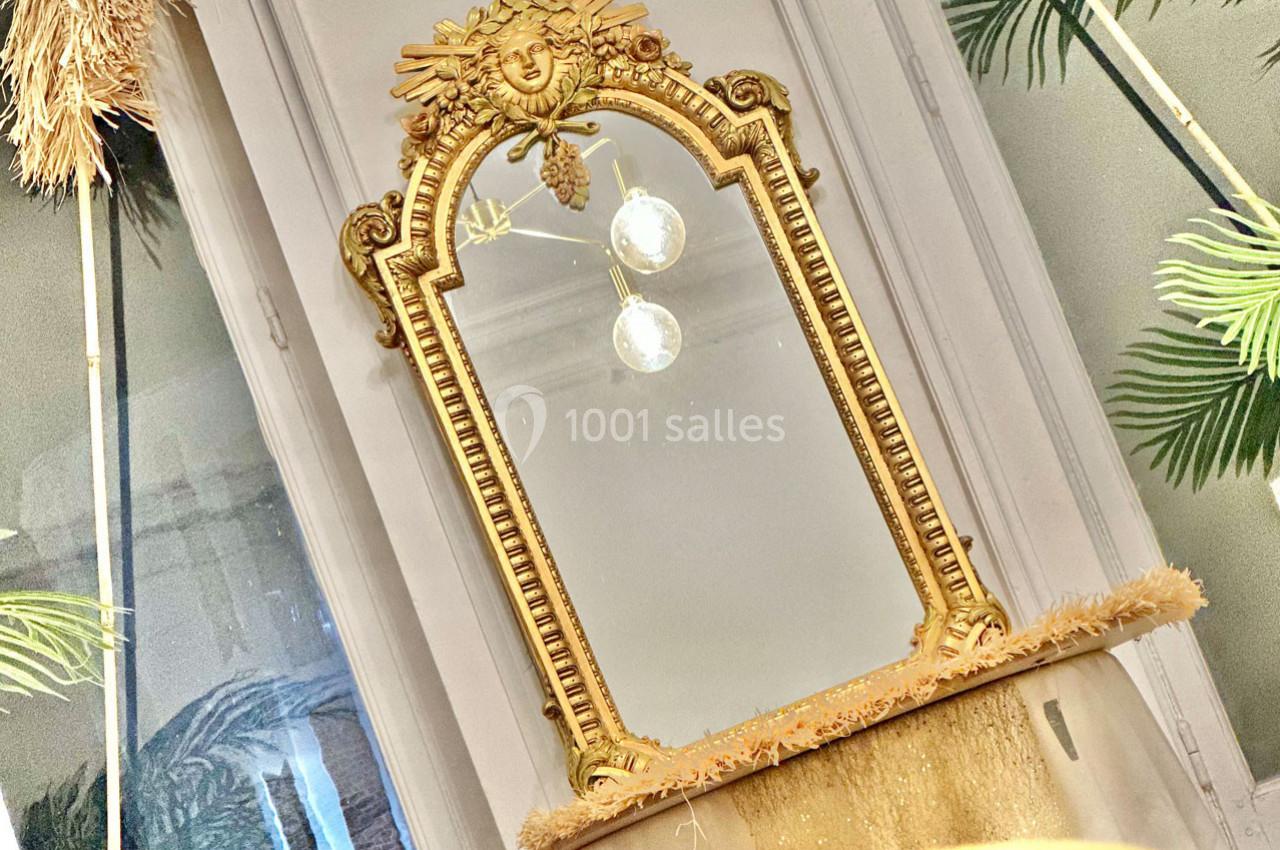 Miroir doré orné de motifs sculptés, posé devant une fenêtre avec des plantes en arrière-plan.