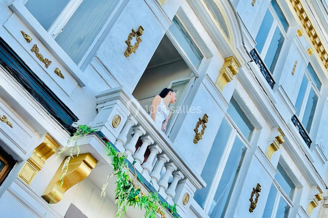 Femme en robe blanche debout sur un balcon orné de végétation, devant une façade blanche et dorée.