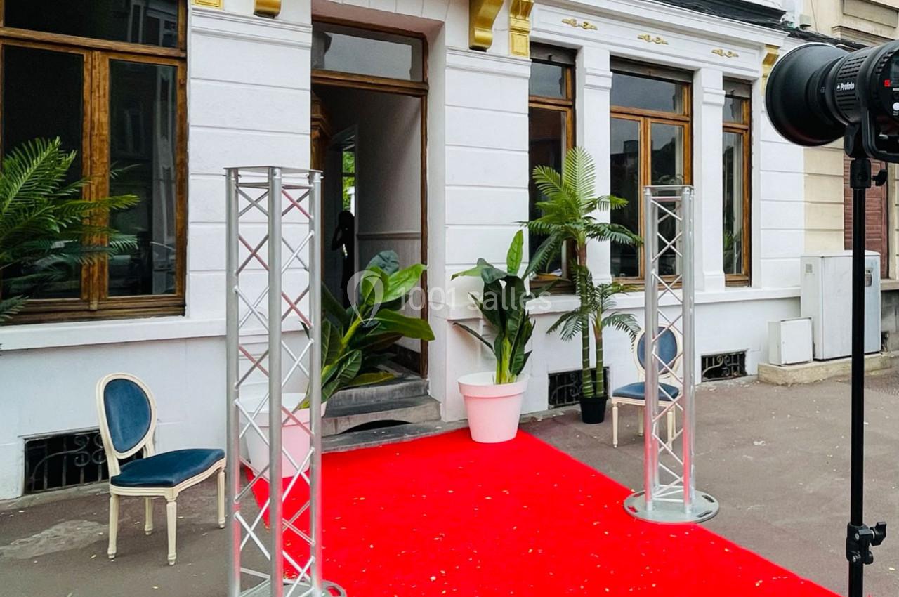 Entrée d'un bâtiment avec tapis rouge, plantes en pot, structures métalliques et chaise sur le côté.