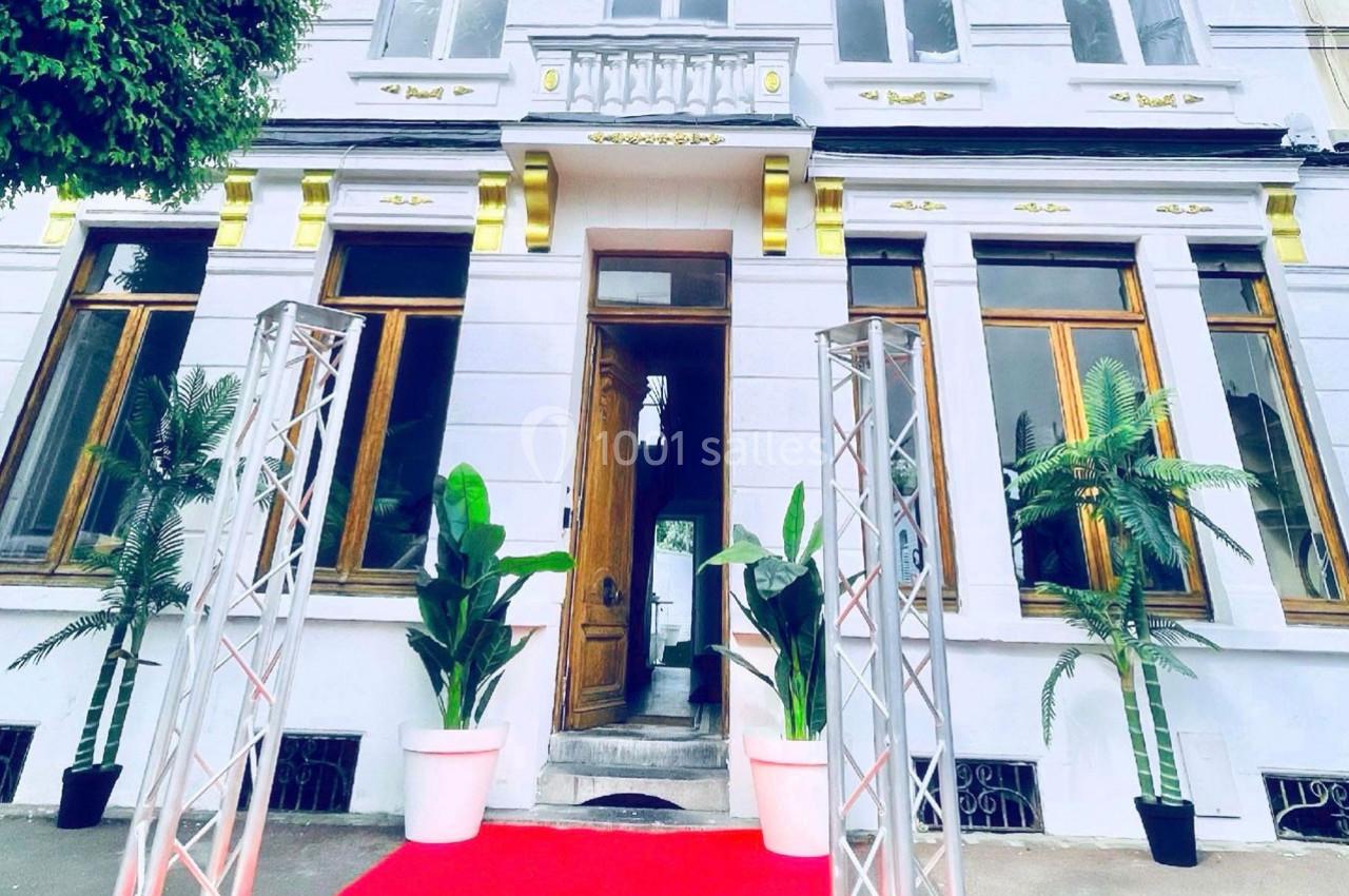 Façade d'un bâtiment blanc avec des fenêtres en bois, entrée centrale ouverte, tapis rouge et plantes en pots.