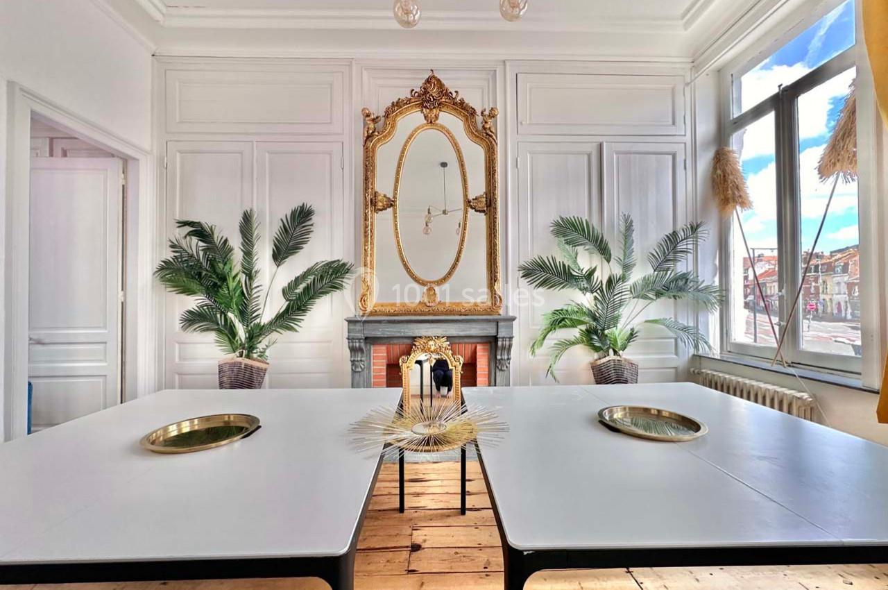 Salle lumineuse avec murs blancs, miroir doré, plantes décoratives, deux tables blanches et vue sur l'extérieur.