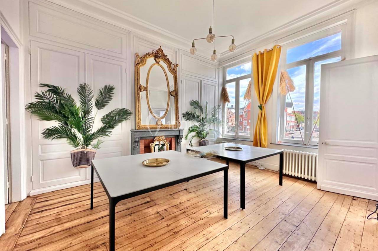 Salle lumineuse avec parquet en bois, tables modernes, miroir doré, plantes et grandes fenêtres avec rideaux jaunes.