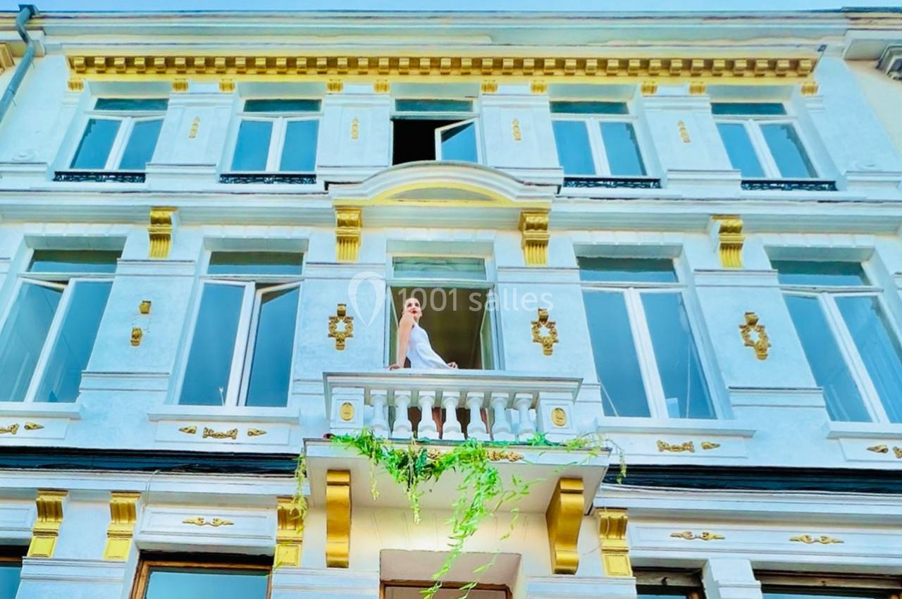 Femme en robe blanche debout sur un balcon orné de plantes, devant une façade élégante aux détails dorés.