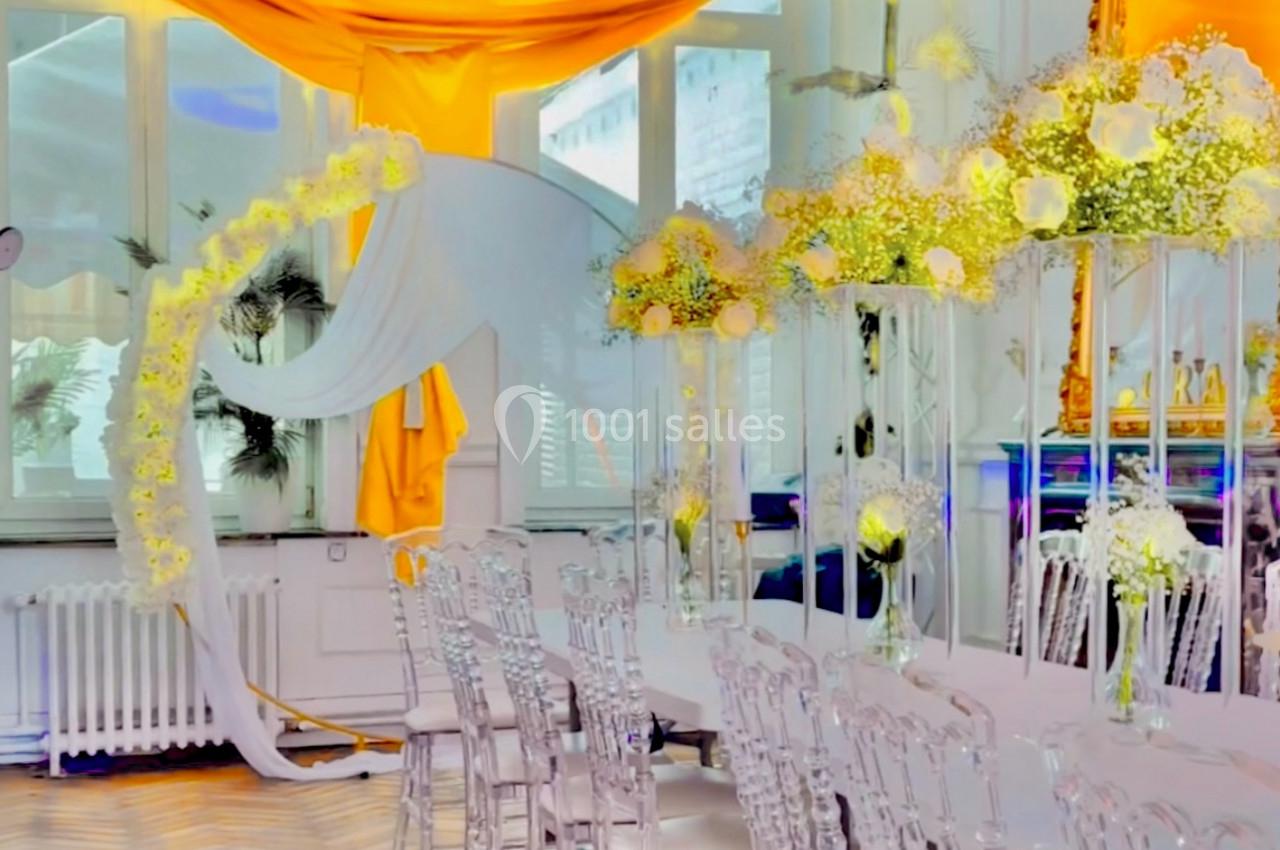Salle décorée avec des chaises transparentes, une table blanche, des fleurs blanches et jaunes, et un drapé orange vif.