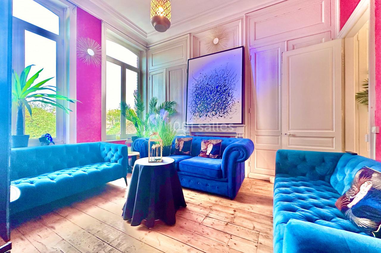 Salon lumineux avec murs roses, canapés bleus, table ronde noire, plantes et tableau abstrait sur un mur blanc.