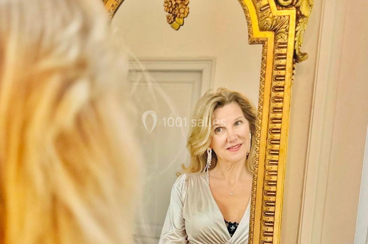 Femme blonde regardant son reflet dans un miroir doré orné de motifs sculptés.
