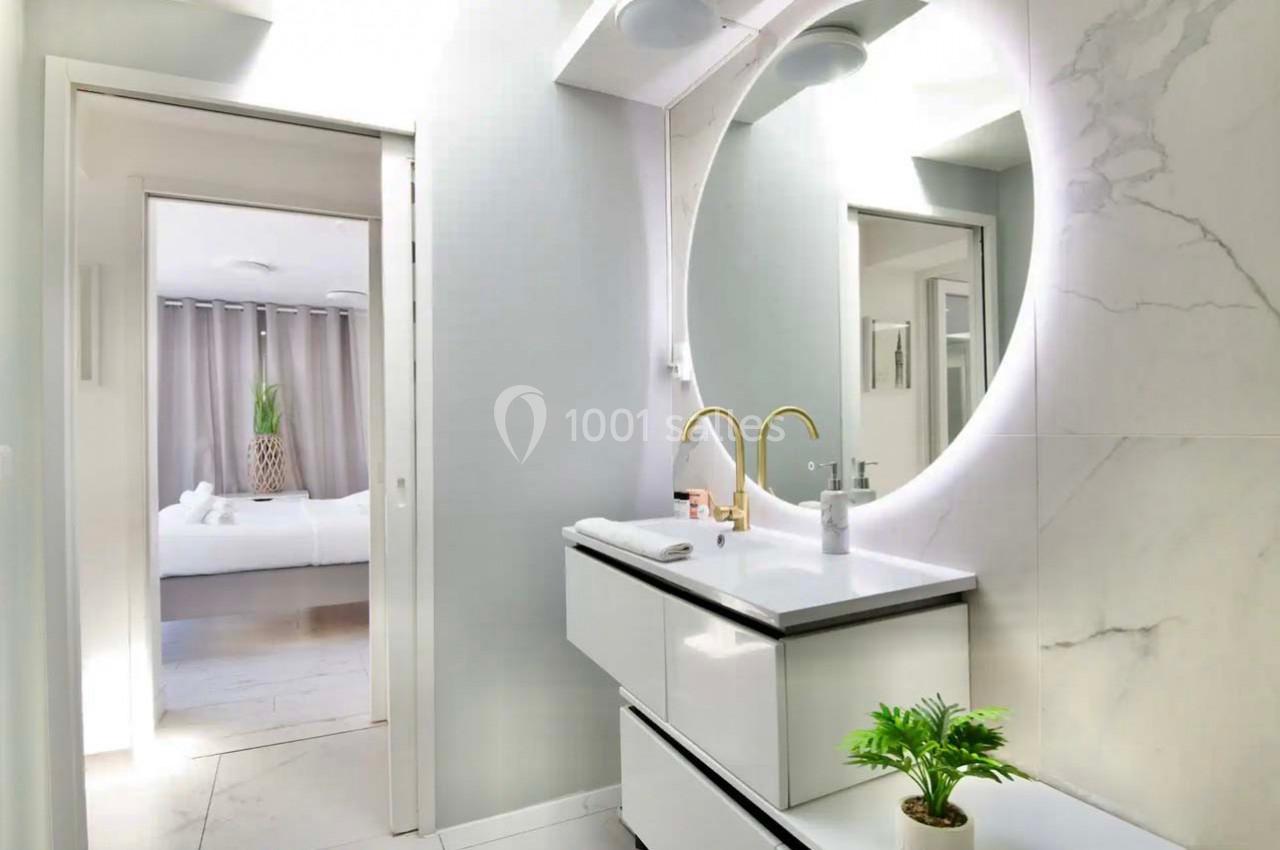 Salle de bain moderne avec lavabo blanc, miroir éclairé circulaire et vue sur une chambre lumineuse en arrière-plan.
